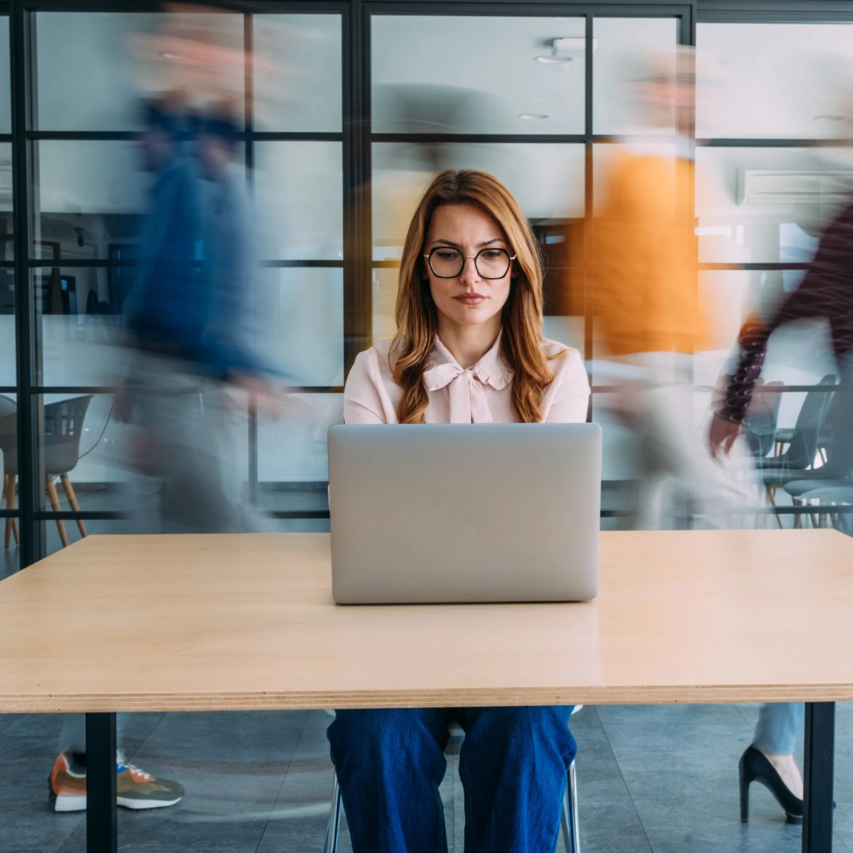 Shot of businesswoman sitting on desk in her office and using laptop. Blurred motion of energetic businesspeople on the move and businesswoman working on laptop in modern office. Creative business person using laptop in the workplace while her colleagues are walking in blurred motion. Busy hours in a board room during working hours while contemporary businesswoman is looking at laptop and her colleagues are passing behind her in a conference room during business hours. Beautiful smiling businesswoman sitting on desk in a busy office, coworkers walk behind her in blurred motion. Shot of a female creative with her colleagues blurred in the background. Woman working at a creative office using her computer and people moving at the background.